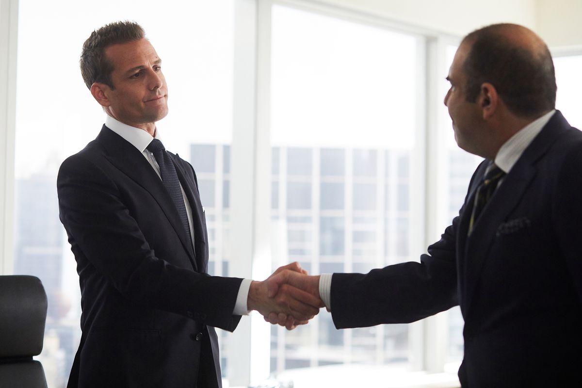 One of the main guys from Suits shakes hands with another guy wearing a suit in the Suits office