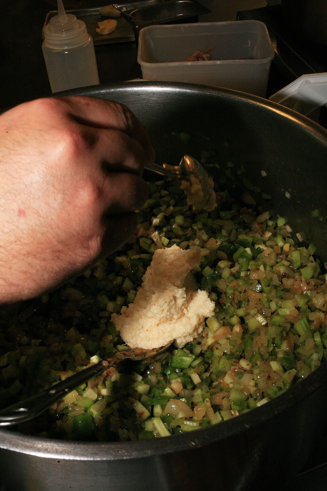 A chef spoons a beige, grated garlic paste into the peppers, celery, and onions, which are now sweated down in the steel pan.
