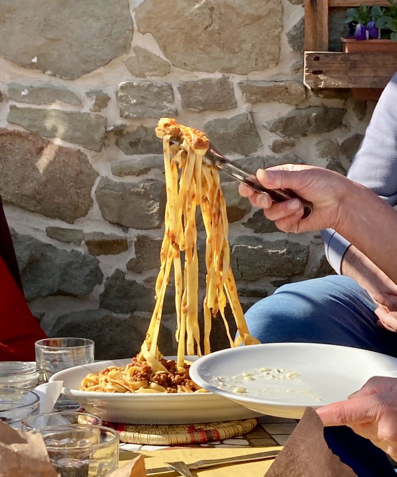 A hand lifts a forkful of pasta from a plate on a sunny table. 
