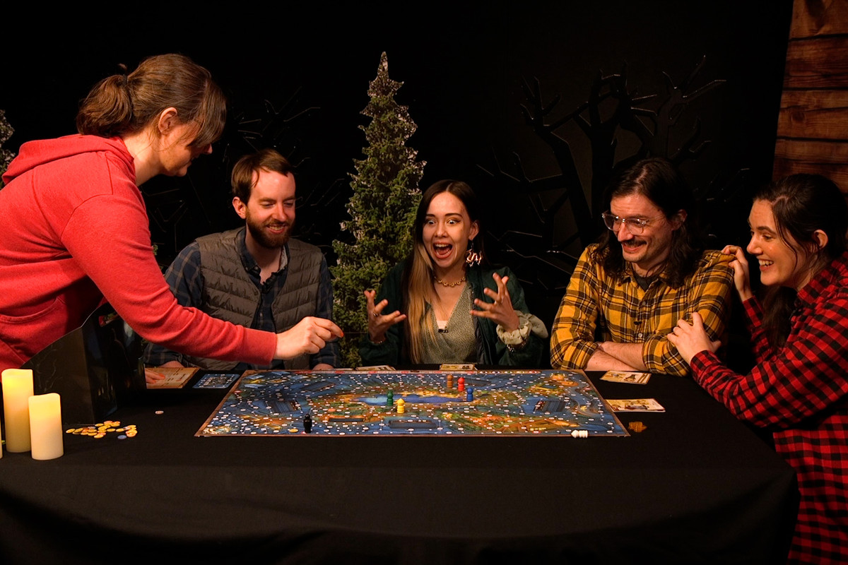A clip from a video where five people are gathered around a table with a large board game. The background set looks like a forest with cardboard cut out trees and a faux cabin wall on the right side. The player at center of the table has an excited and terrified look on their face as the player on the left prepares to place something on the board. The rest of the players look on with intense trepidation.