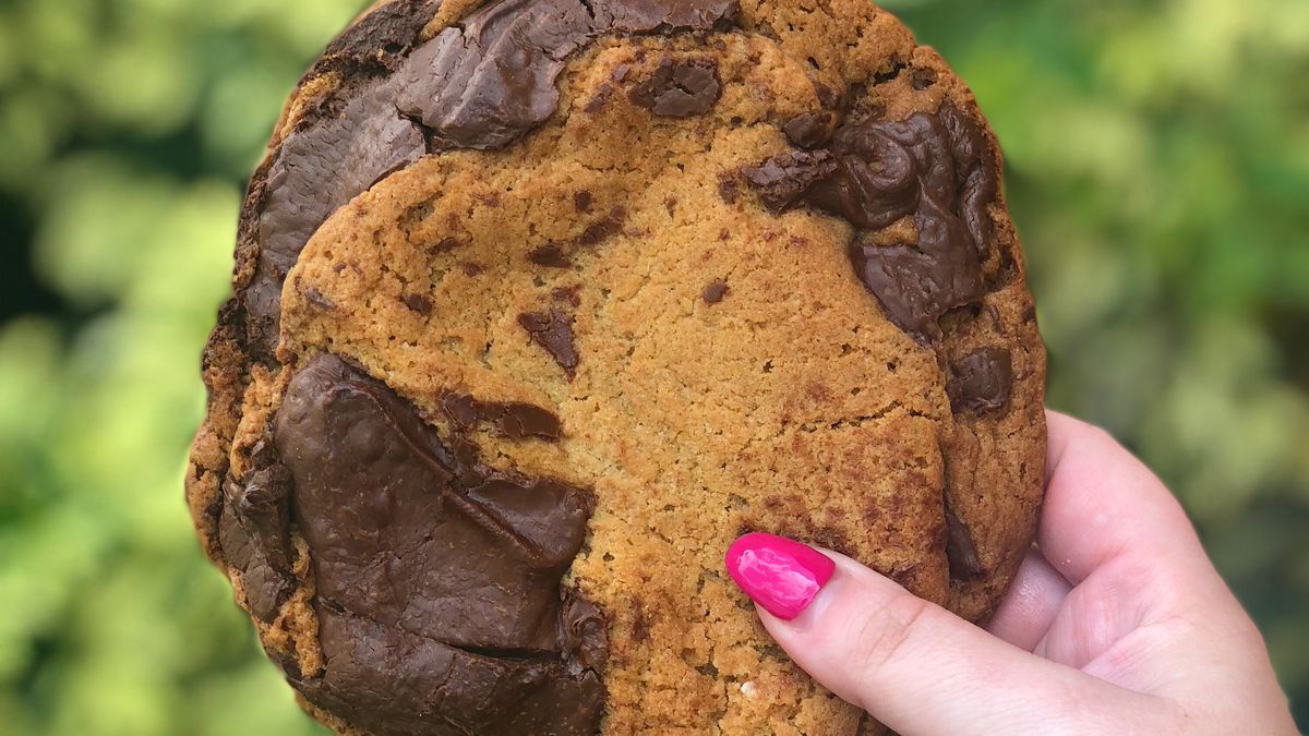 A white hand with pink manicured nails holds up a cookie with huge chunks of melty chocolate chips. 