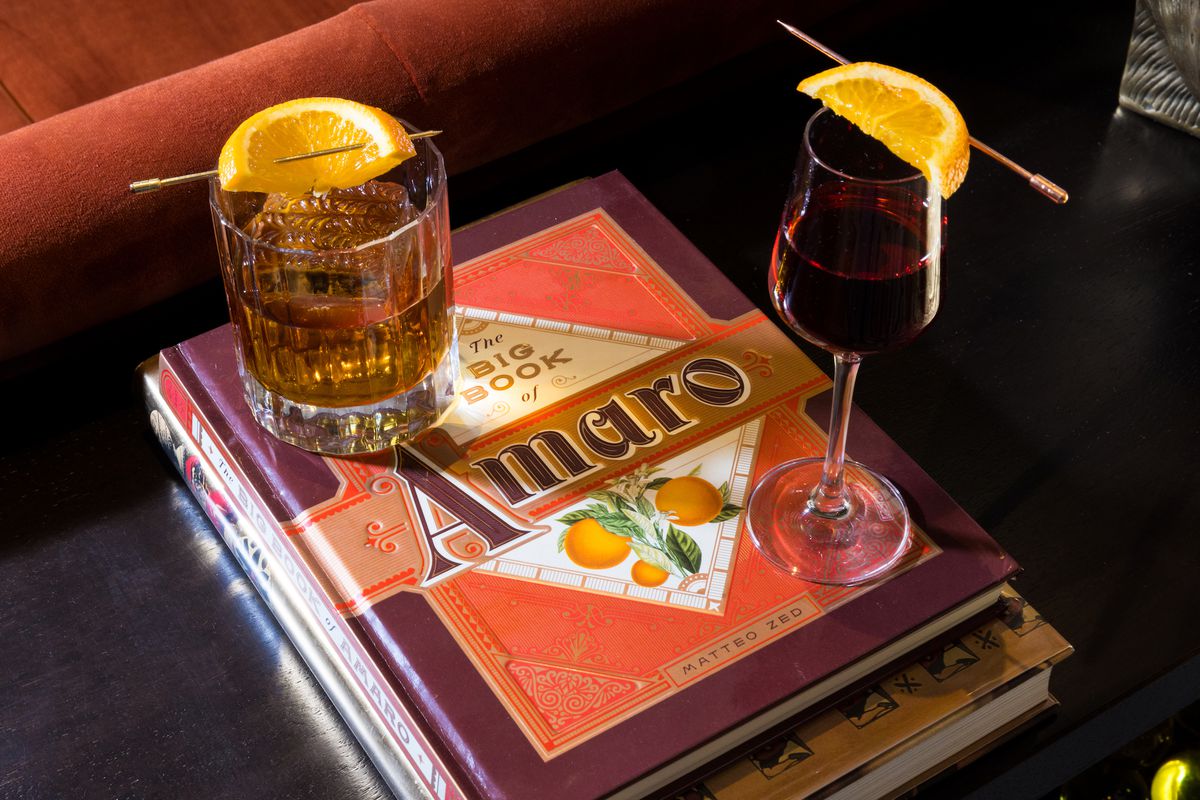 A red-hued image with drinks and orange slices on a book about Amaro.