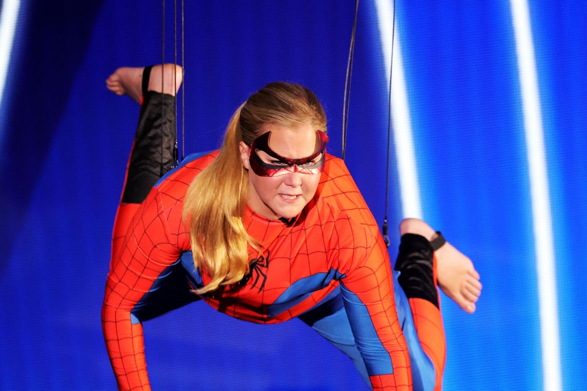 Amy Schumer is dressed as Spider-Man and hanging from wires in front of a blue backdrop on a stage. 