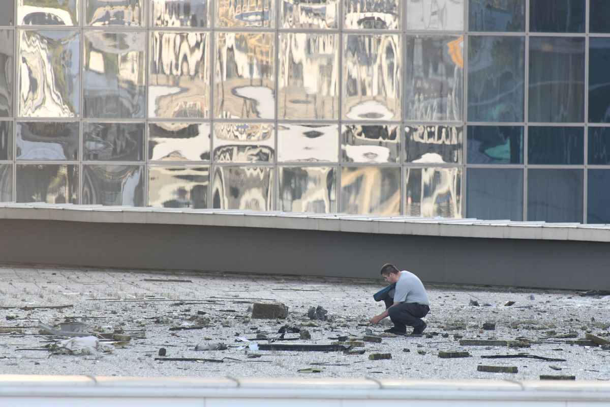 A man crouches on the sidewalk to inspect rubble and debris in front of a mirrored wall.