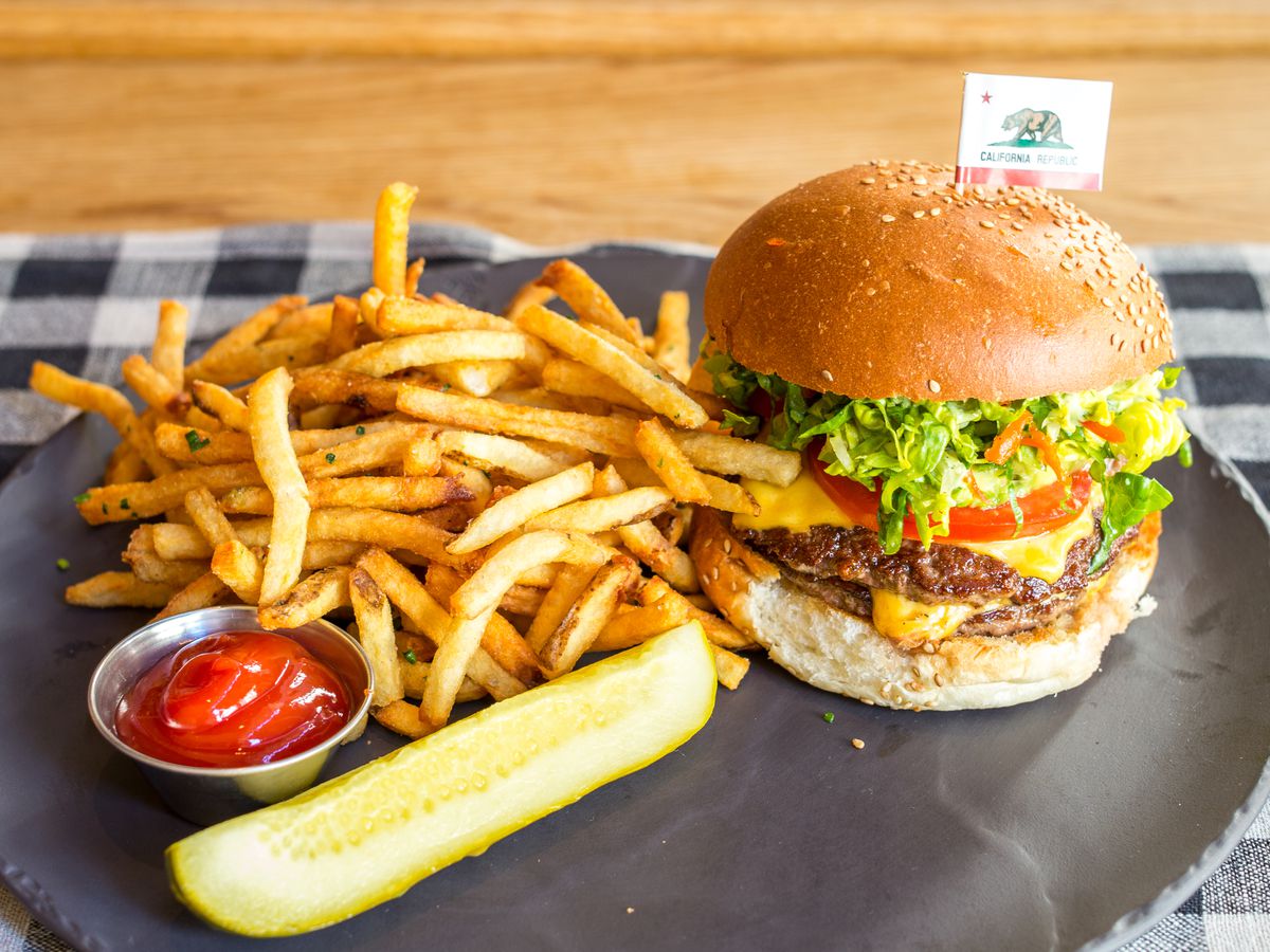 A side of thinly-cut french fries, ketchup, a quarter pickle, and a sesame seed burger with the works (lettuce, tomato, onion, pickle, and cheese) sit on a ceramic plate