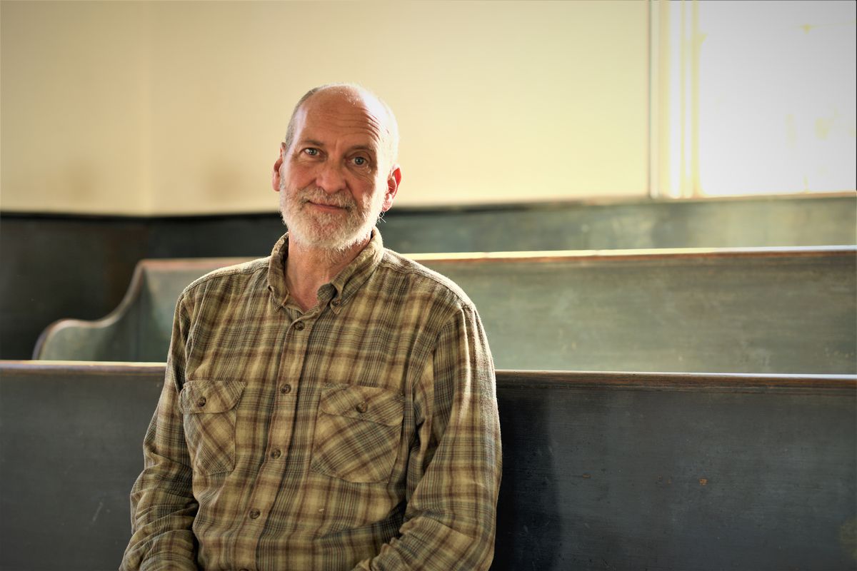An elderly white man in a plaid shirt sits in a church pew, looking at the camera.
