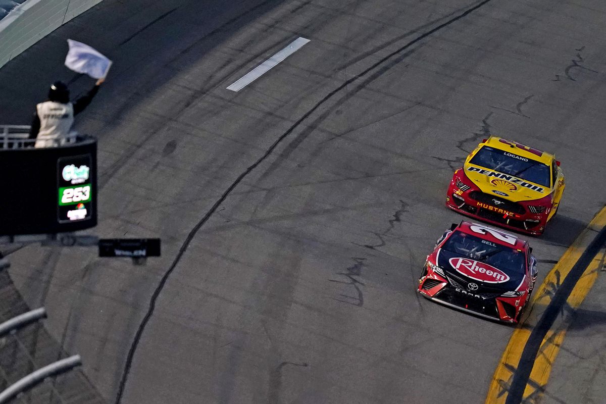 Two NASCAR stock cars approach a finish line with the final lap white flag flying in the forground