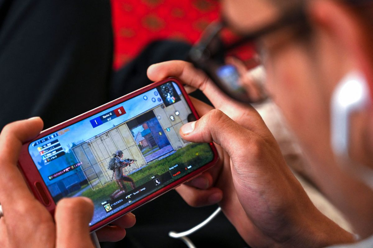 A photograph of a person playing PlayerUnknown’s Battlegrounds (PUBG) on a mobile phone. The camera is pointed at the phone’s screen from behind, and we only see the player’s hands.