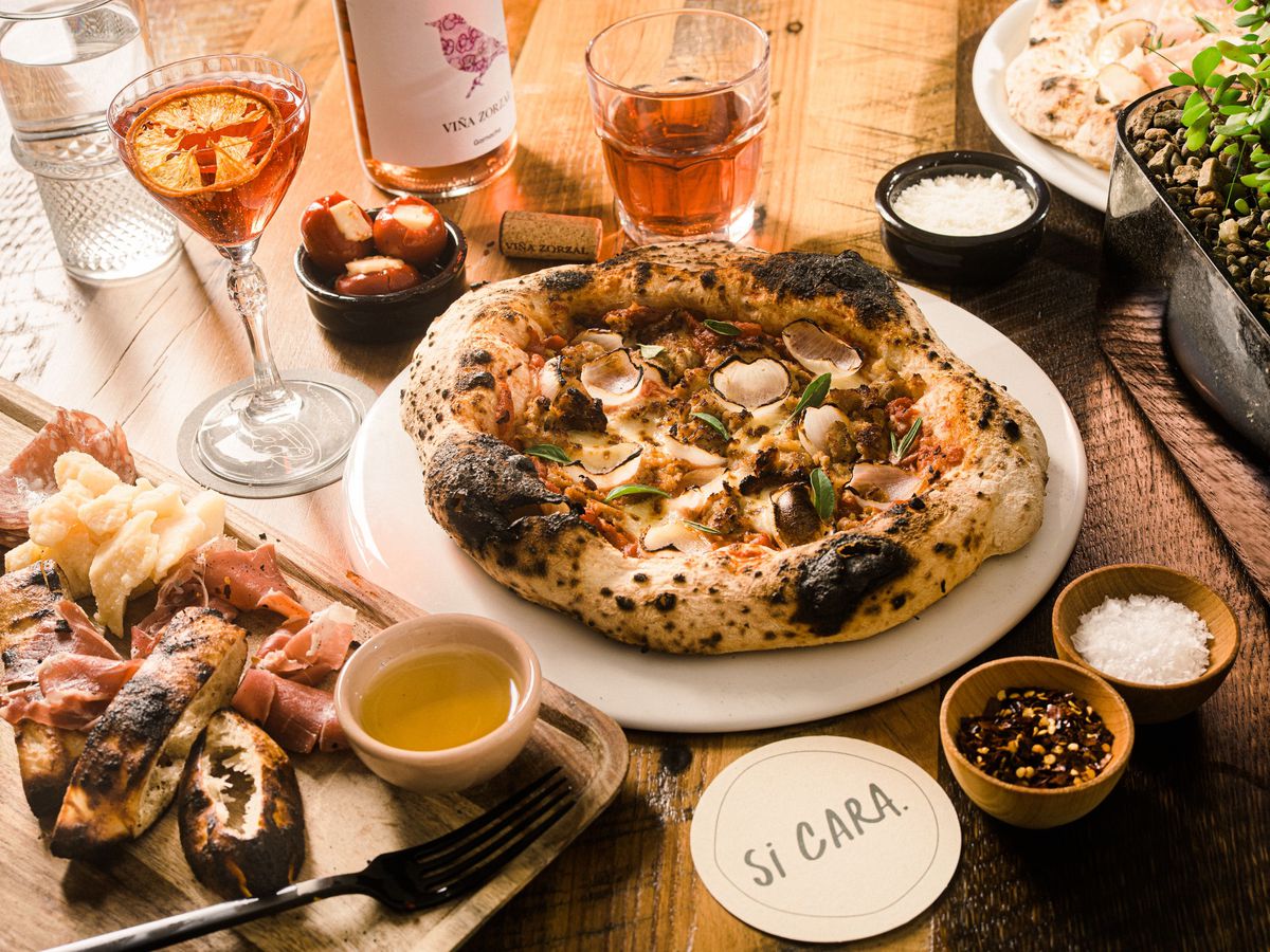 A Neapolitan-ish pizza with an extra-puffy, charred crust sits on a plate on a wooden table, surrounding by glasses of wine; a platter of bread, meat, and cheese; and more.