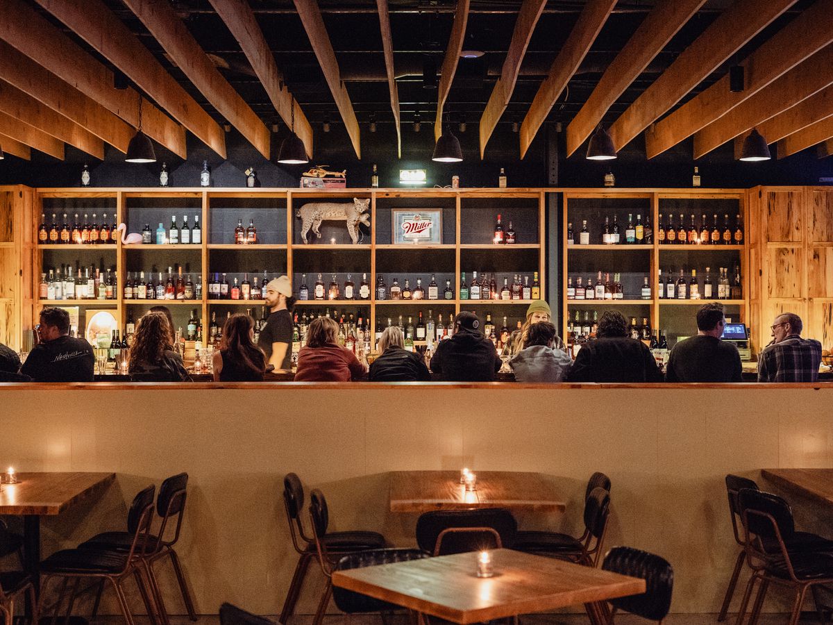 A long stretch of bar with wood paneling on the wall and on the ceiling. Shelves stocked with whiskey line the back of the bar and a row of people are seated at the bar. 