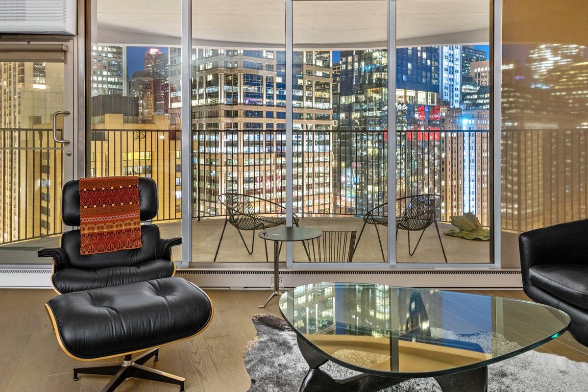 An Eames chair and Noguchi coffee table in a living room with floor to ceiling glass overlooking a balcony and a row of tall skyscrapers at night. 