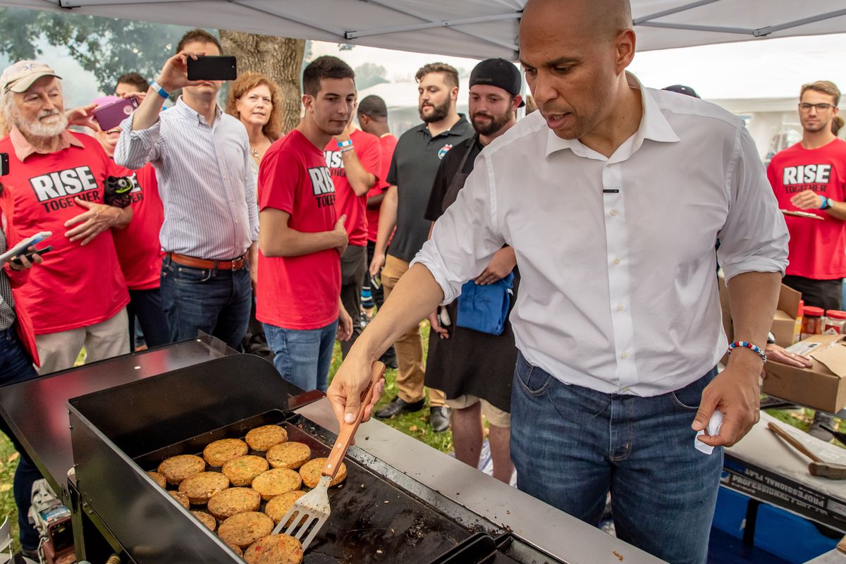 Cory Booker flips over a hamburger bun on a grill with a spatula.