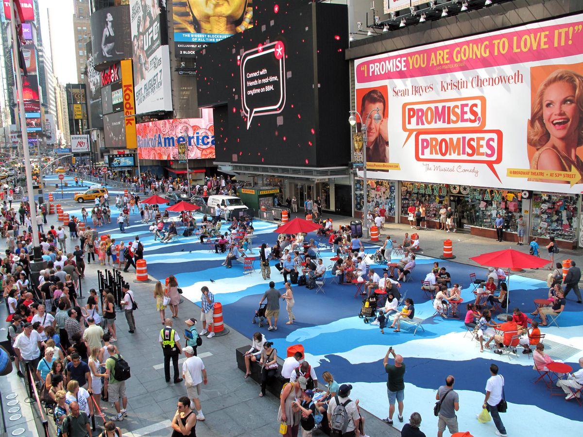 A busy New York City street hung with Broadway posters is painted with a rippling blue mural where people are sitting on beach chairs and under umbrellas.
