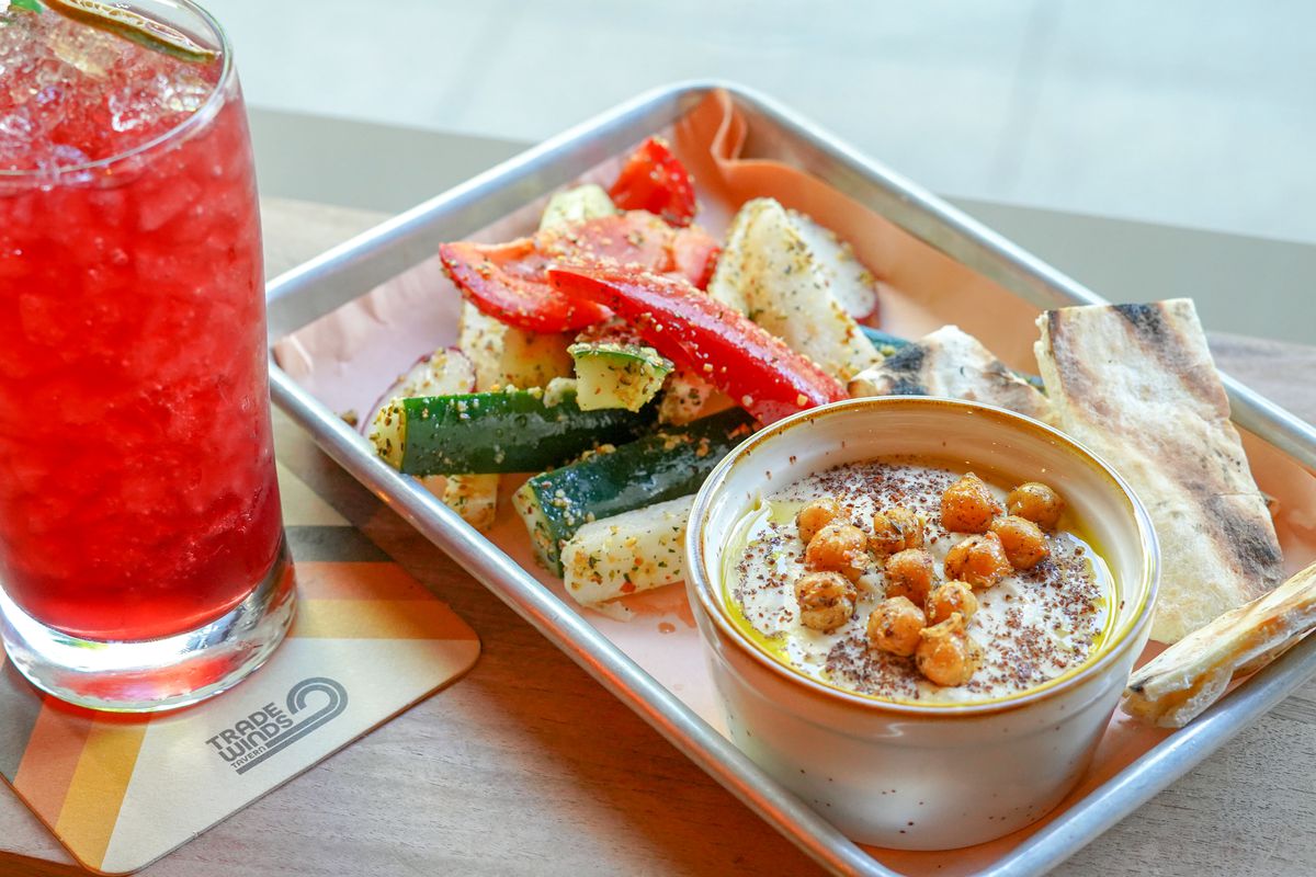 A red drink with ice called the Cape Codder, with vodka, cranberry, pomegranate molasses, and lime oil, alongside a hummus plate with seasoned vegetables and a side of pita