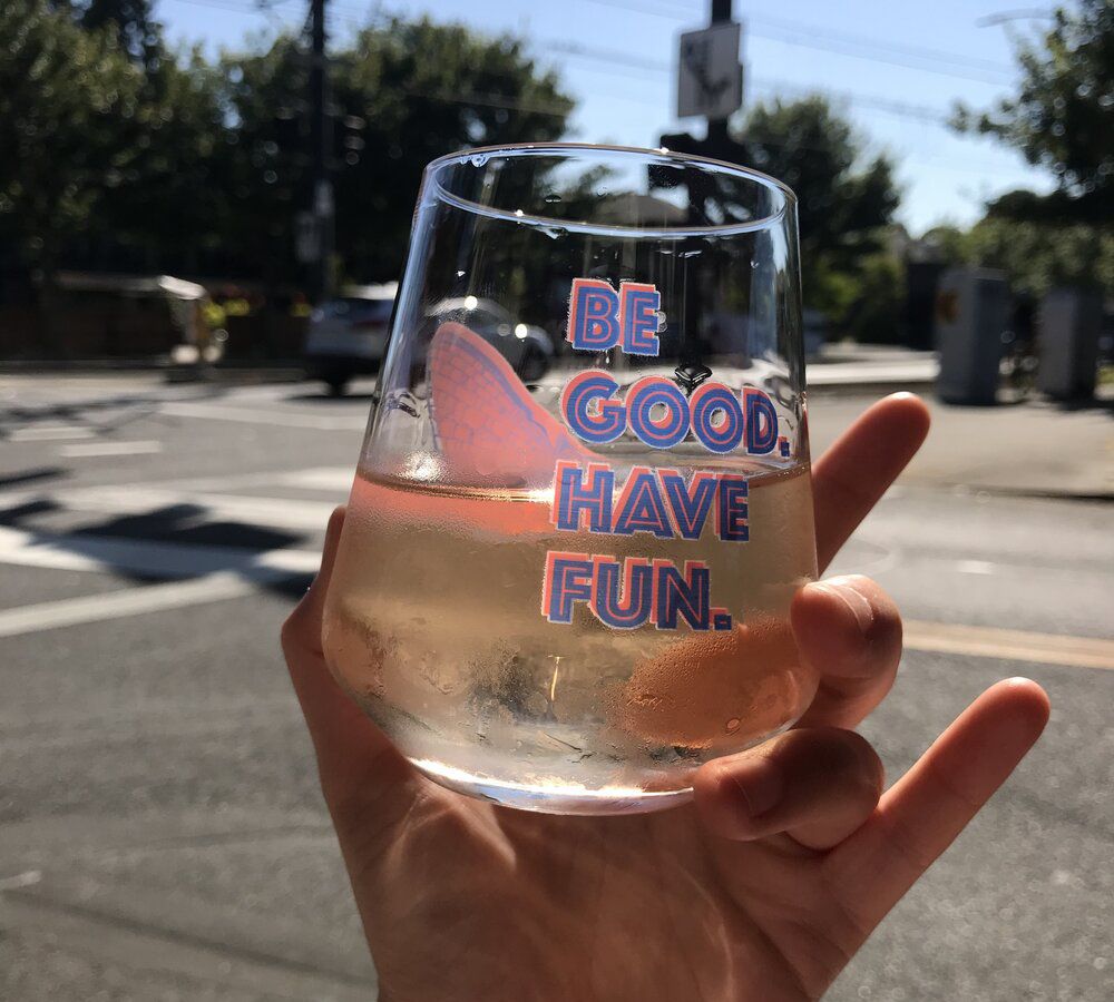 A hand holds a beverage in a glass reading “Be Good Have Fun” on a sunny street corner.