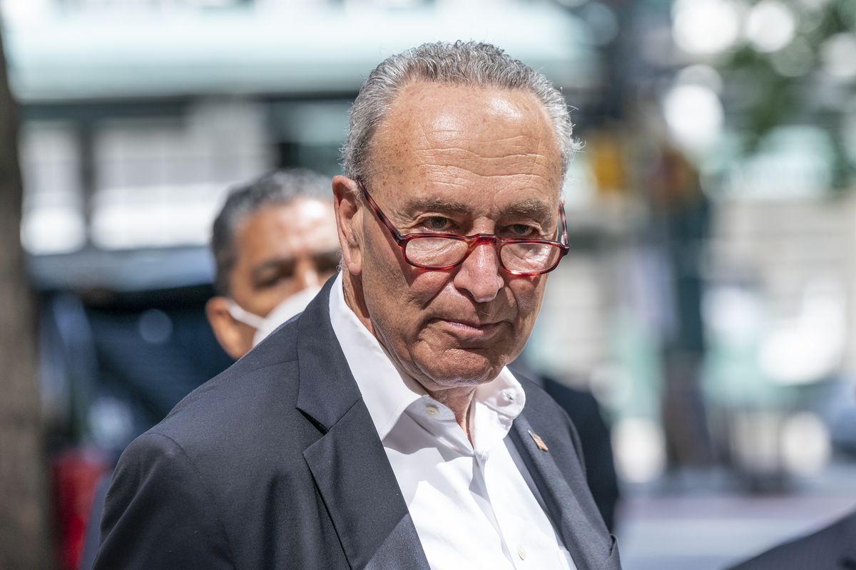Schumer, his red glasses perched on his nose, looks at the camera gravely, standing at a podium in a grey suit and open collared white shirt.