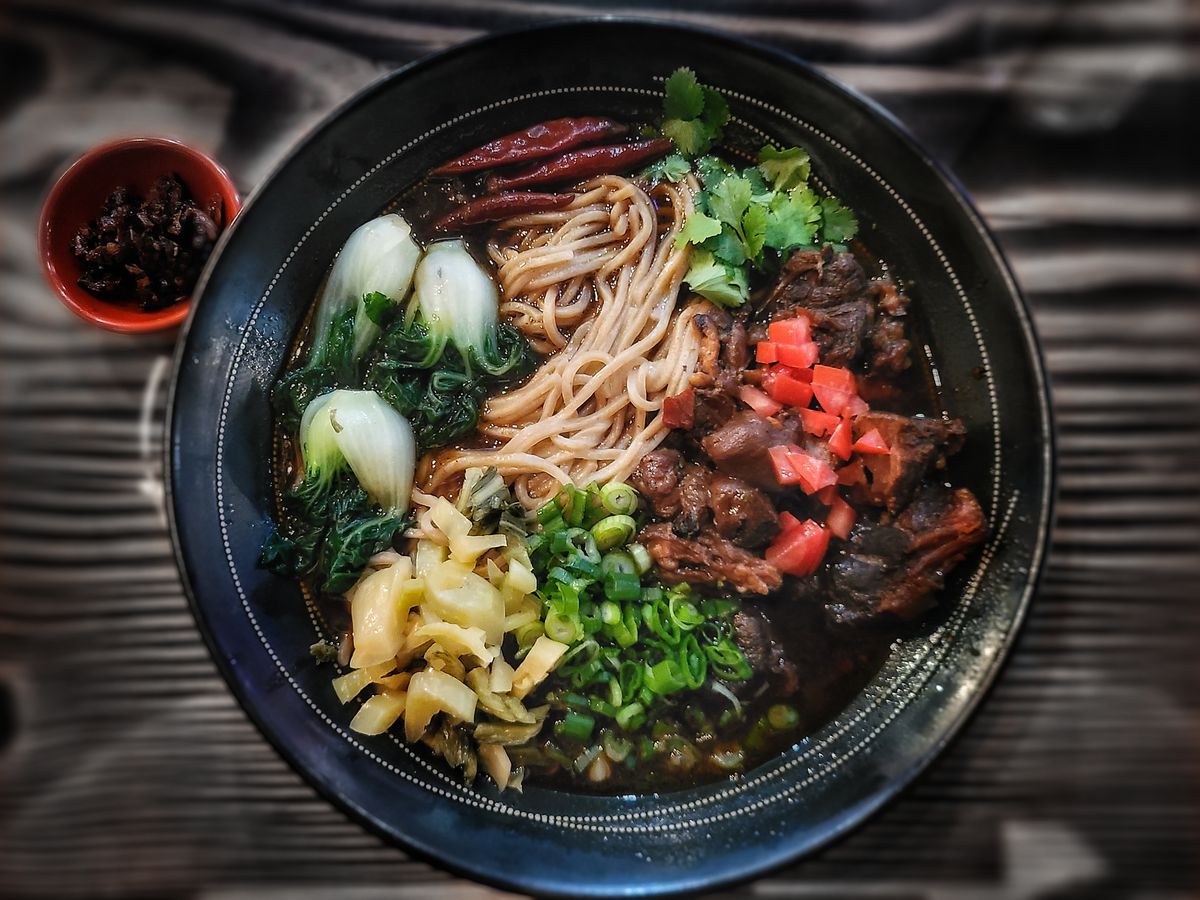 A full, dark colored ramen bowl is shown, including noodles, tomatoes, yu choy, green onion, beef, and cilantro.