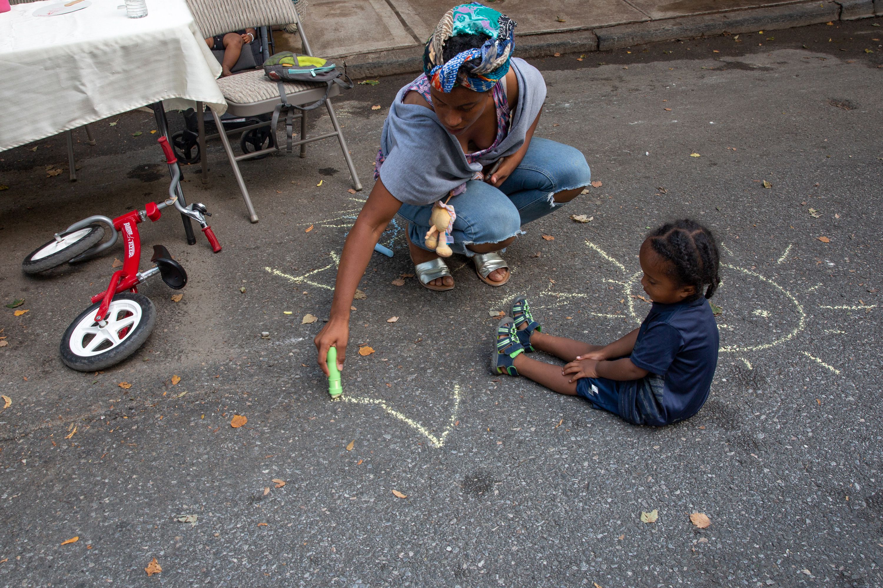 A mother draws on the sidewalk with chalk while her child watches.