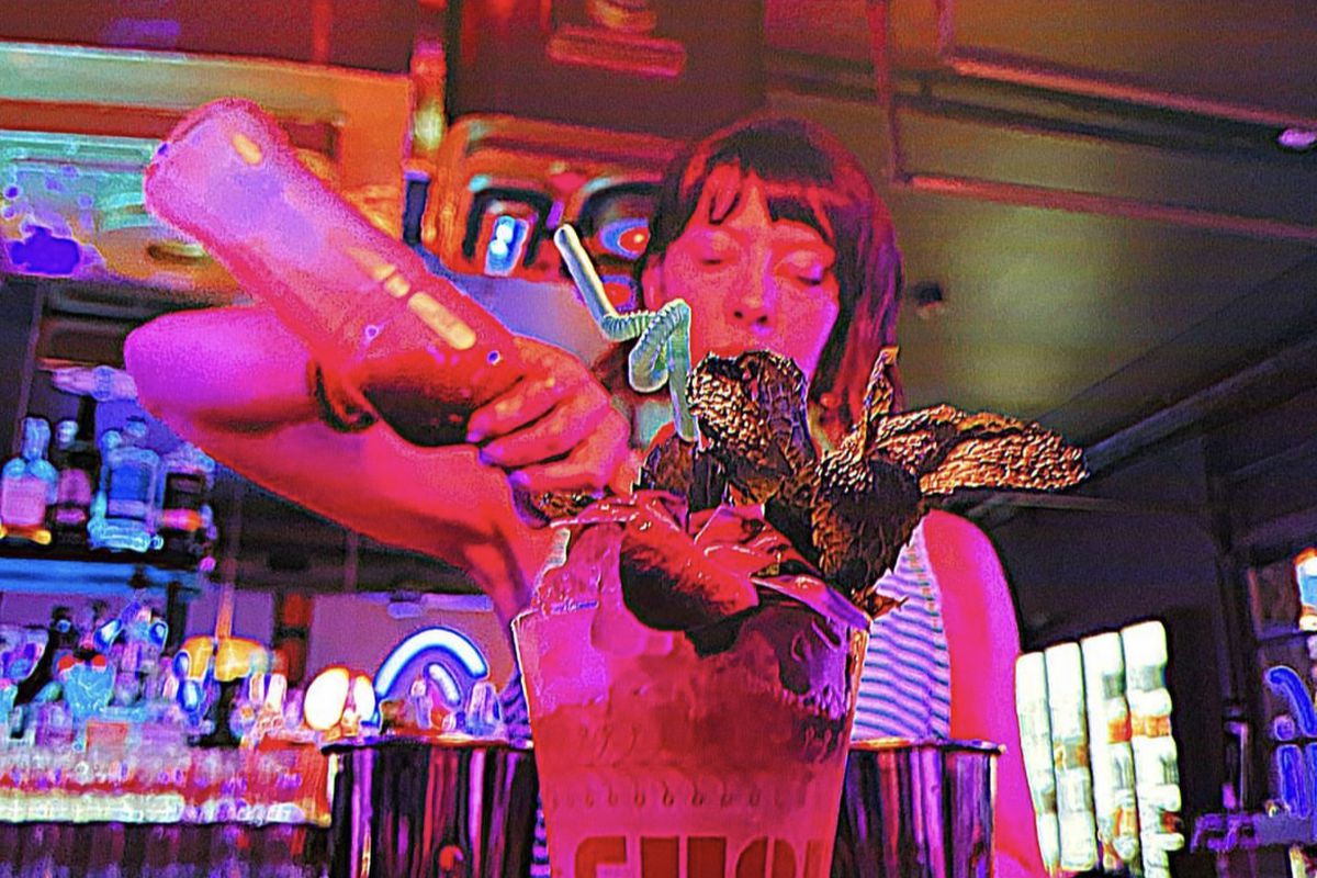 A woman with dark hair and bangs pouring alcohol into a cup with a twisty straw and garnishments.