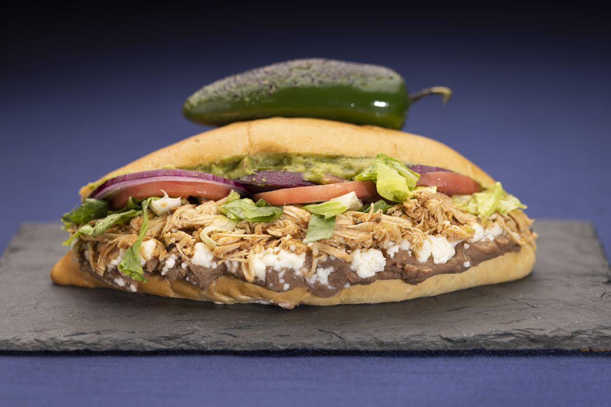 A Bolillo bun is layered with fresh refried beans, choice of house made salsa chicken, barbacoa or pastor pork, then topped with queso fresco, lettuce, tomato, red onion, and guacamole.&nbsp;