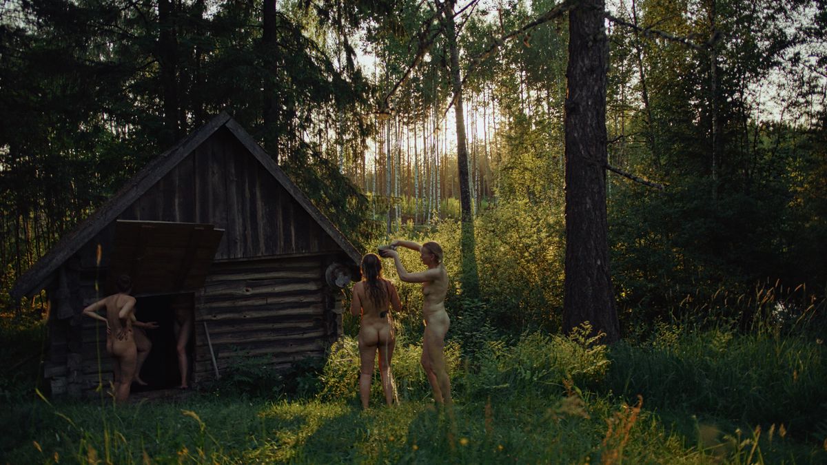 Four nude women stand around or inside a small sauna made of raw logs, standing in a vividly green forest, in Smoke Sauna Sisterhood