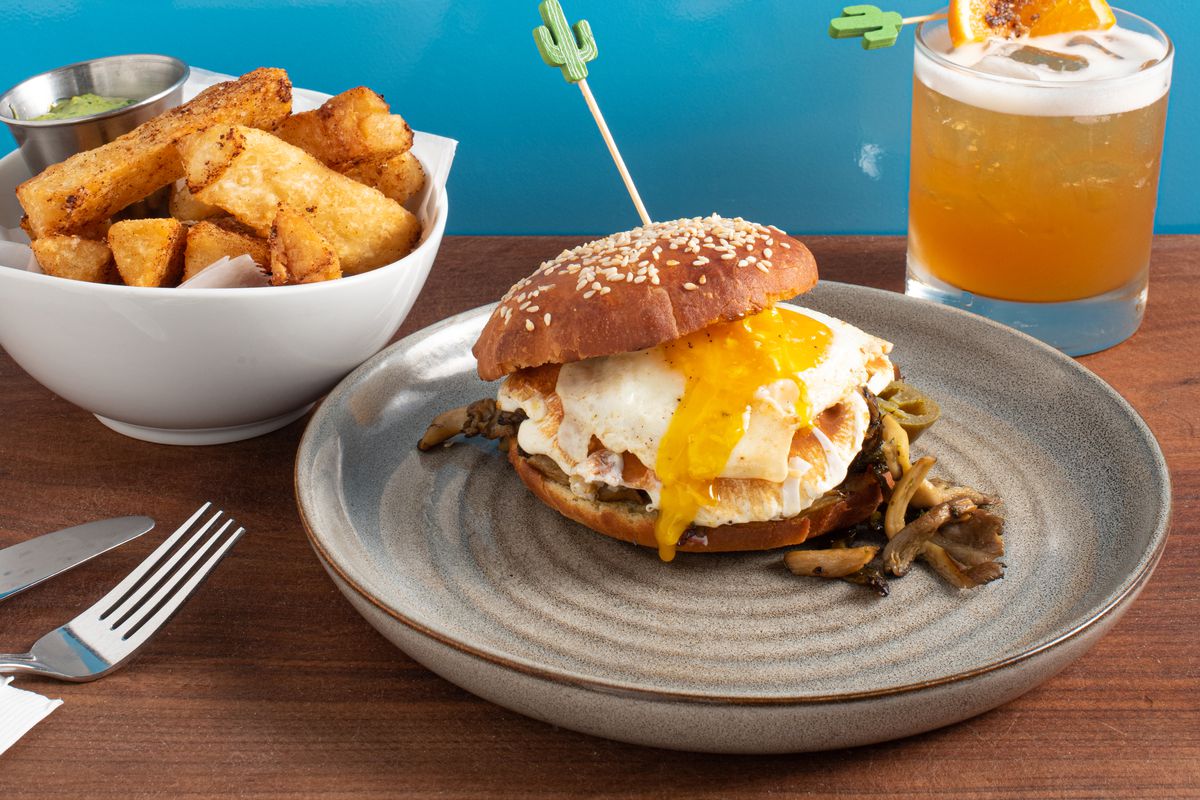 A breakfast sandwich with a gooey egg yolk on a bun is front and center, with an orange cocktail with a frothy top, and a white bowl of crispy potatoes in the background.