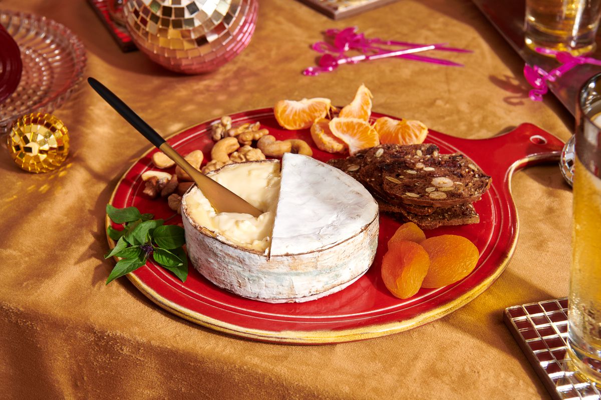 A wheel of cheese sits on a platter with nuts, dried apricots, and crackers.