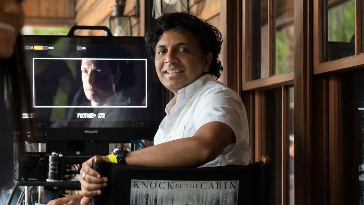 M. Night Shyamalan sitting in his director’s chair and looking back at the camera as footage from Knock at the Cabin is on a screen in front of him.