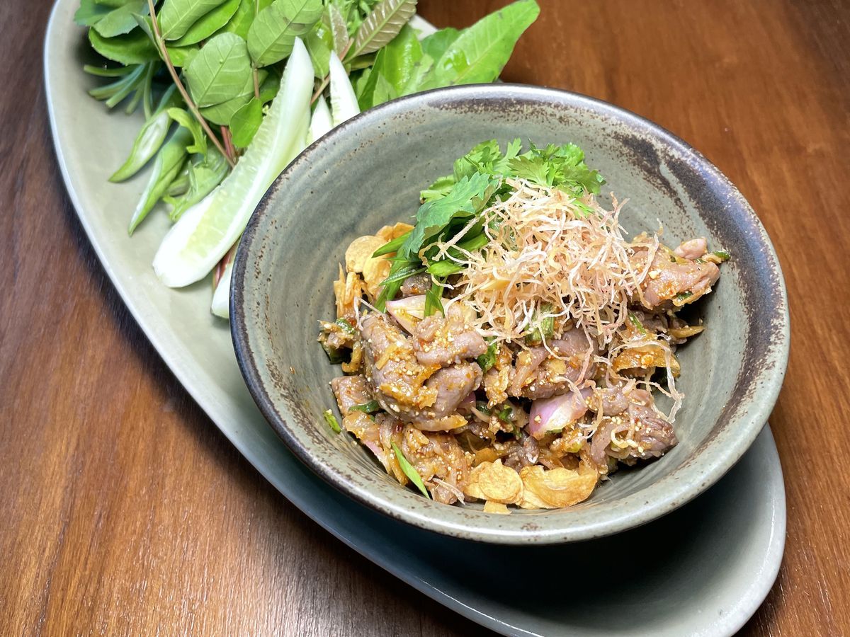 A bowl of duck larb beneath a pile of crispy fixings, presented on a long tray with greens.