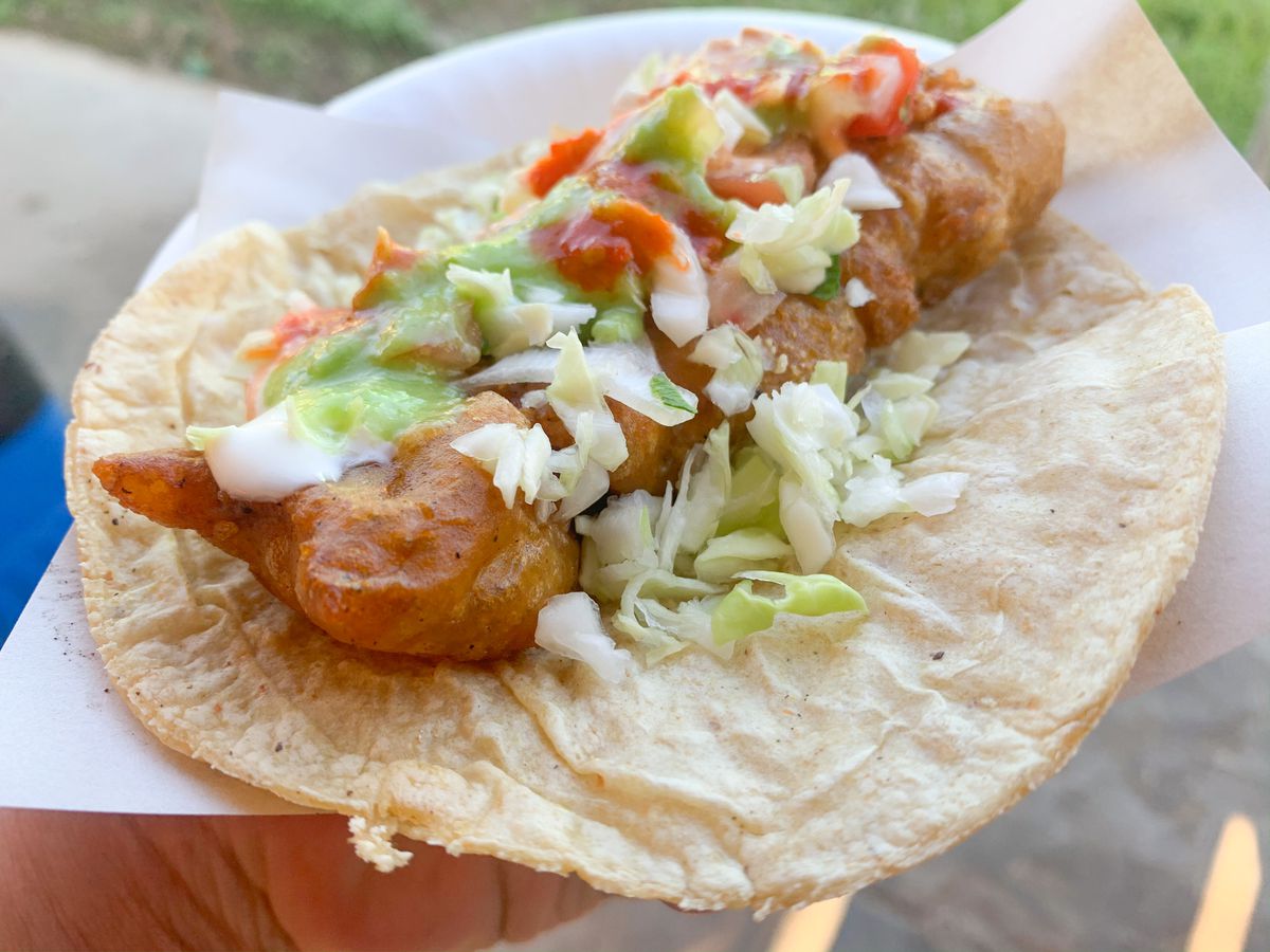 A hand holds a taco outside. A long piece of fried fish sits in the taco topped with lettuce, onions, and sauces