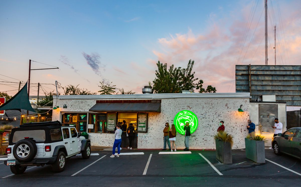 A line of people standing socially distanced and in masks ready to order from Supremo Taco’s to-go window. The shot is taken from the parking lot looking toward the white painted brick building with the bright neon green Supremo logo lit up as the evening sun sets, A white Jeep is parked to the left