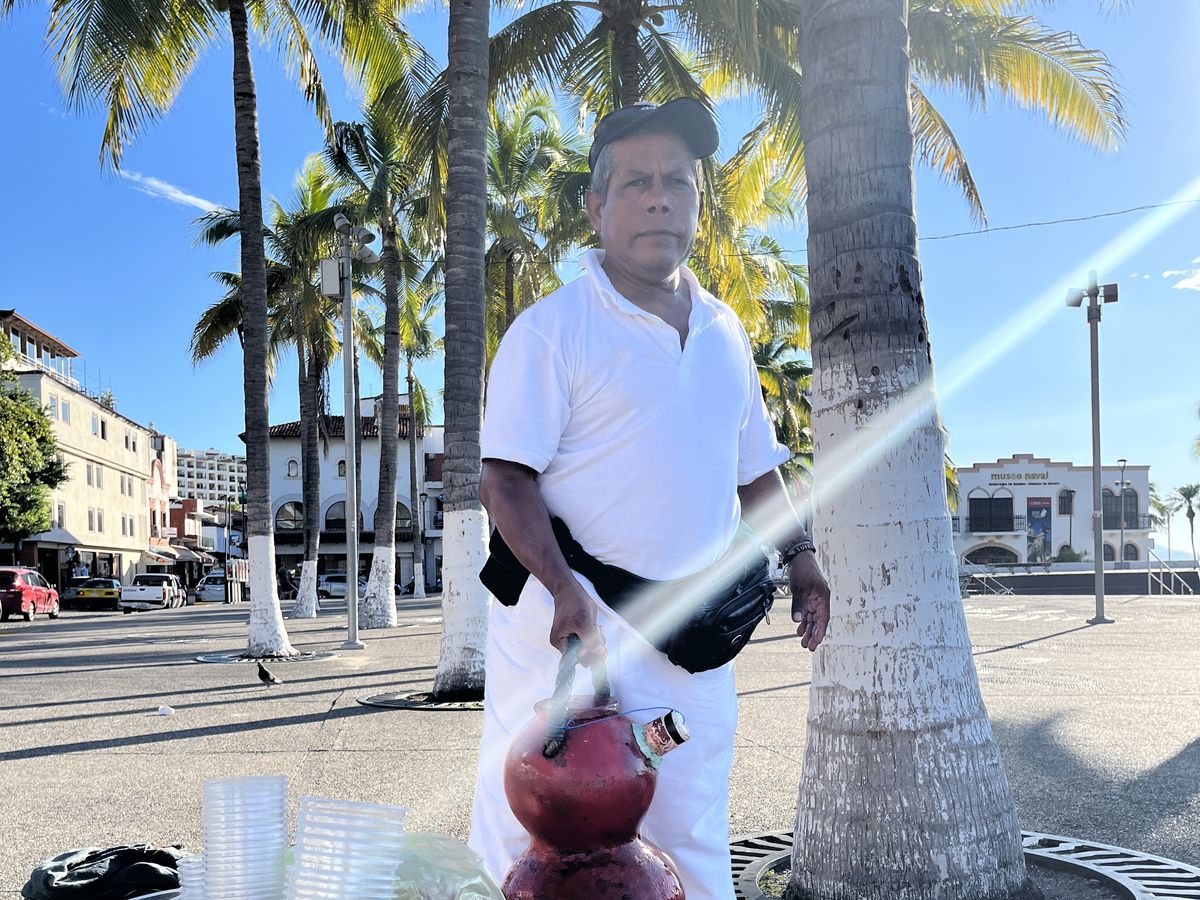 A vendor stands with a jug and other drink supplies on a sunny stretch of concrete between palm trees.