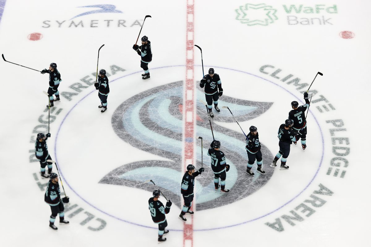 A bird’s-eye shot of the Kraken gathering around the center ice logo as they lift their sticks into the air to thank the fans after their first home win.