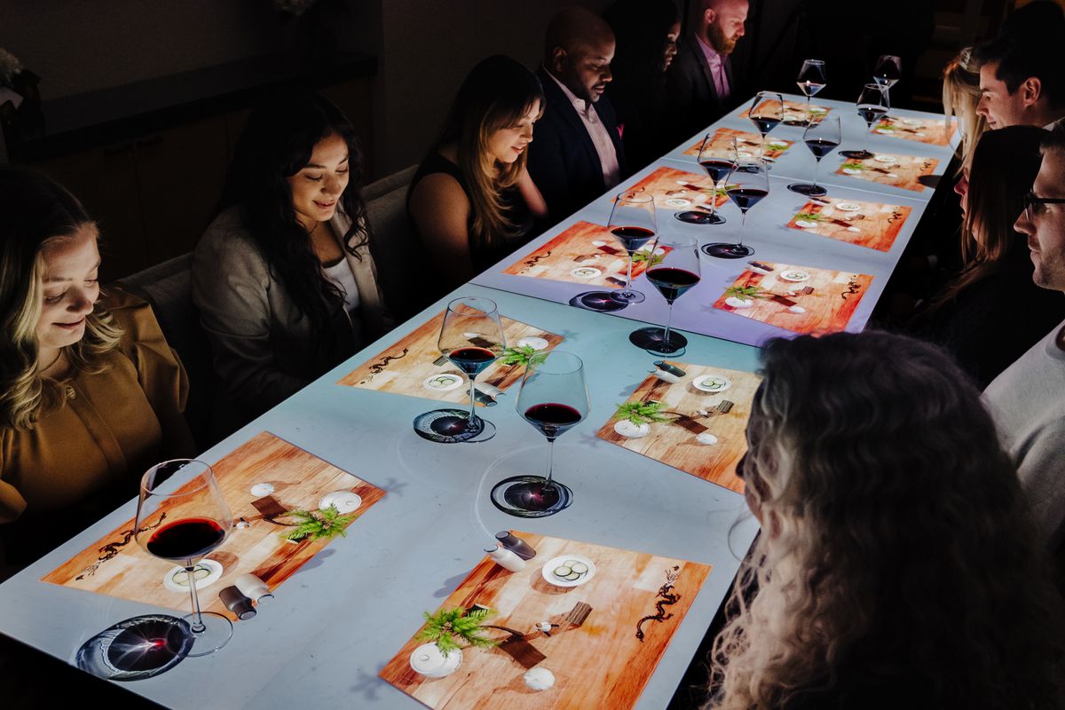 People seated at a long dinner table with individual animations of a plate setup in front of them.