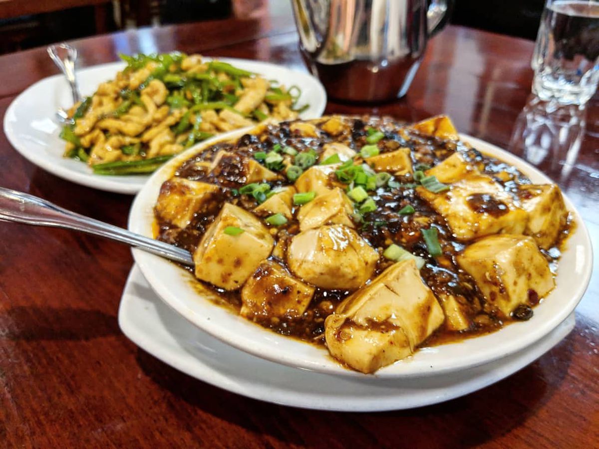 A white bowl on a glossy wooden table is filled to the brim with a meaty rendition of mapo tofu, topped with scallions.