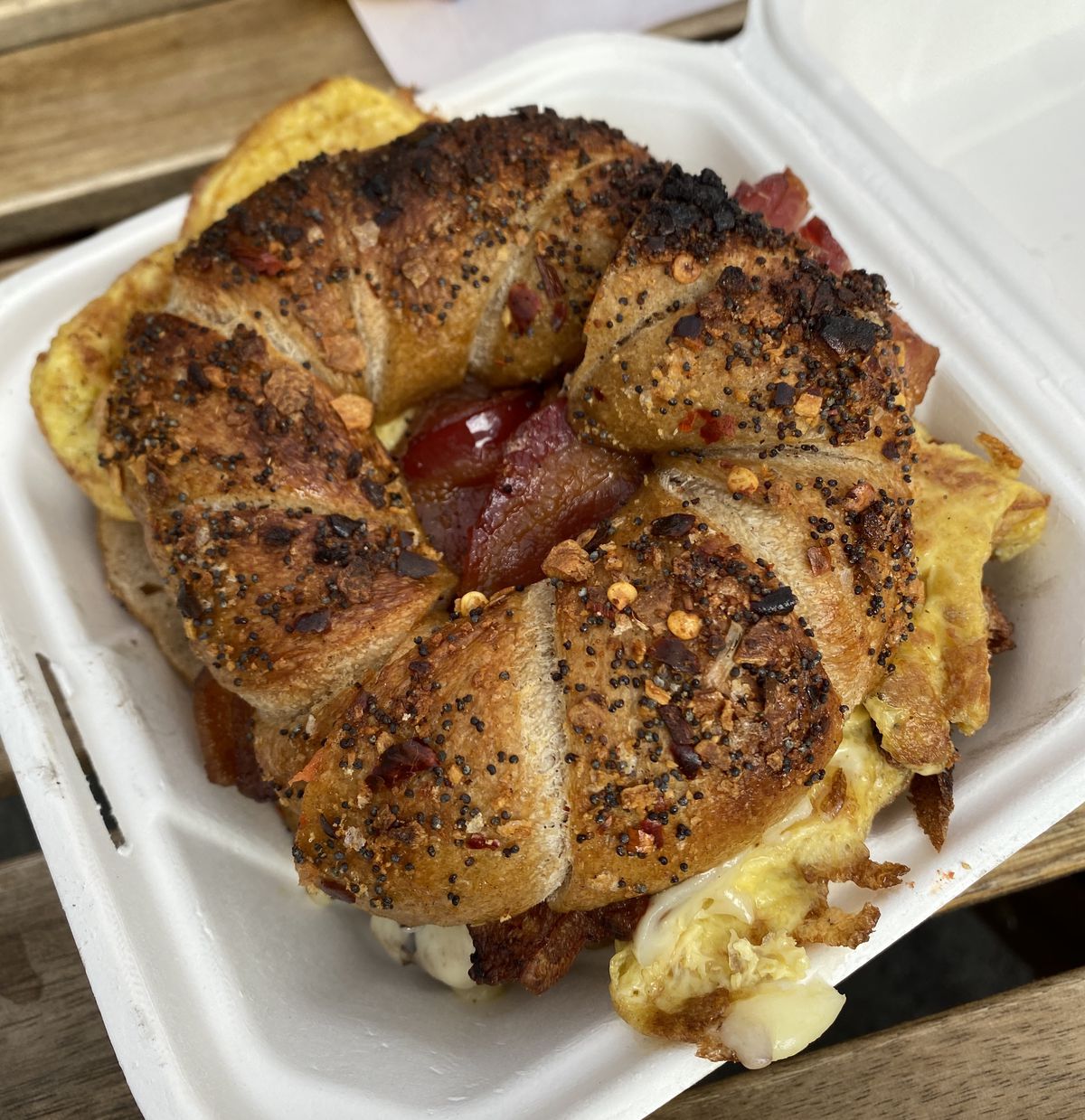 A bacon, egg, and cheese sandwich on a toasted everything bagel with red pepper flakes sits in a cardboard white takeout container