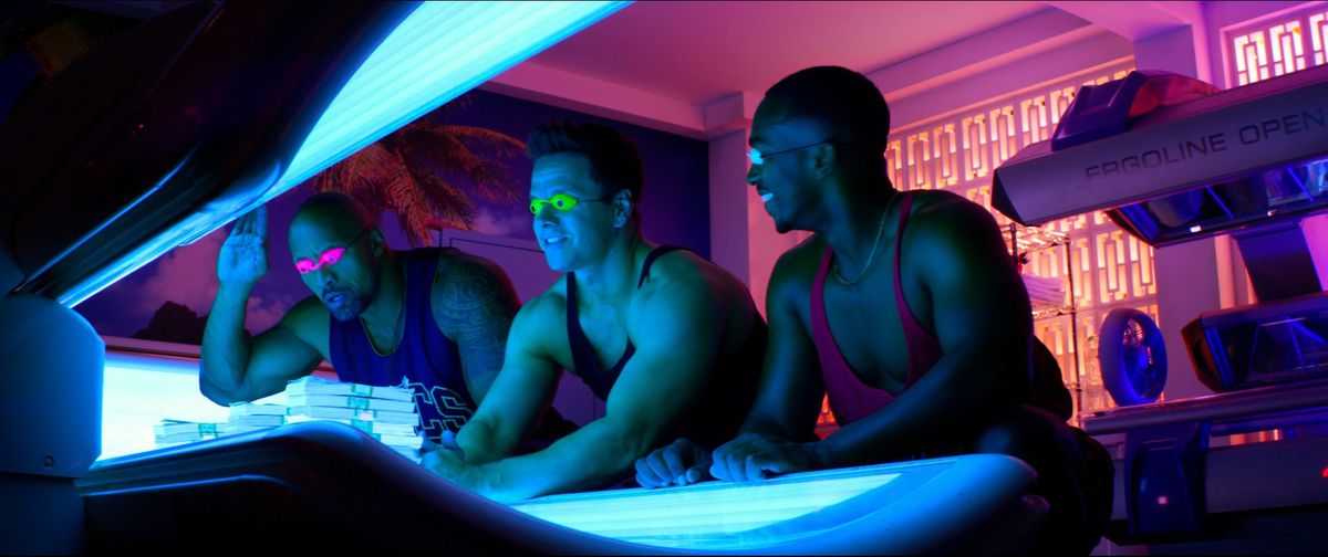 Dwayne Johnson, Mark Wahlberg, and Anthony Mackie smile in front of a tanning bed while all wearing colorful tanktops in Pain and Gain.