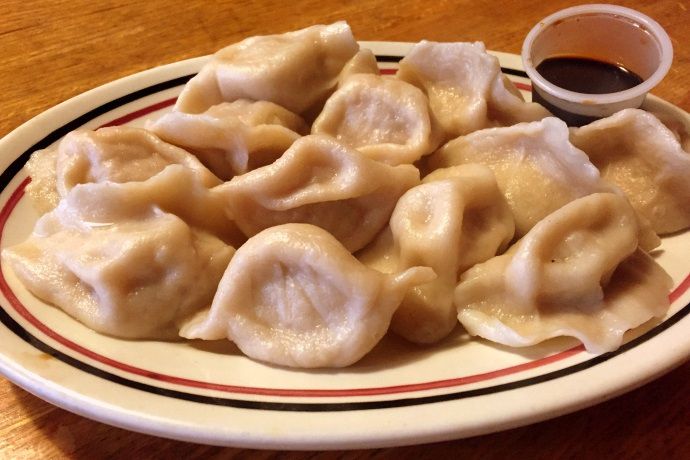 A dozen boiled, plump, Chinese-style dumplings sit on an off-white plate with black and orange circles around the rim. There’s a plastic cup of soy sauce on the plate, too. The plate is on a wooden table.