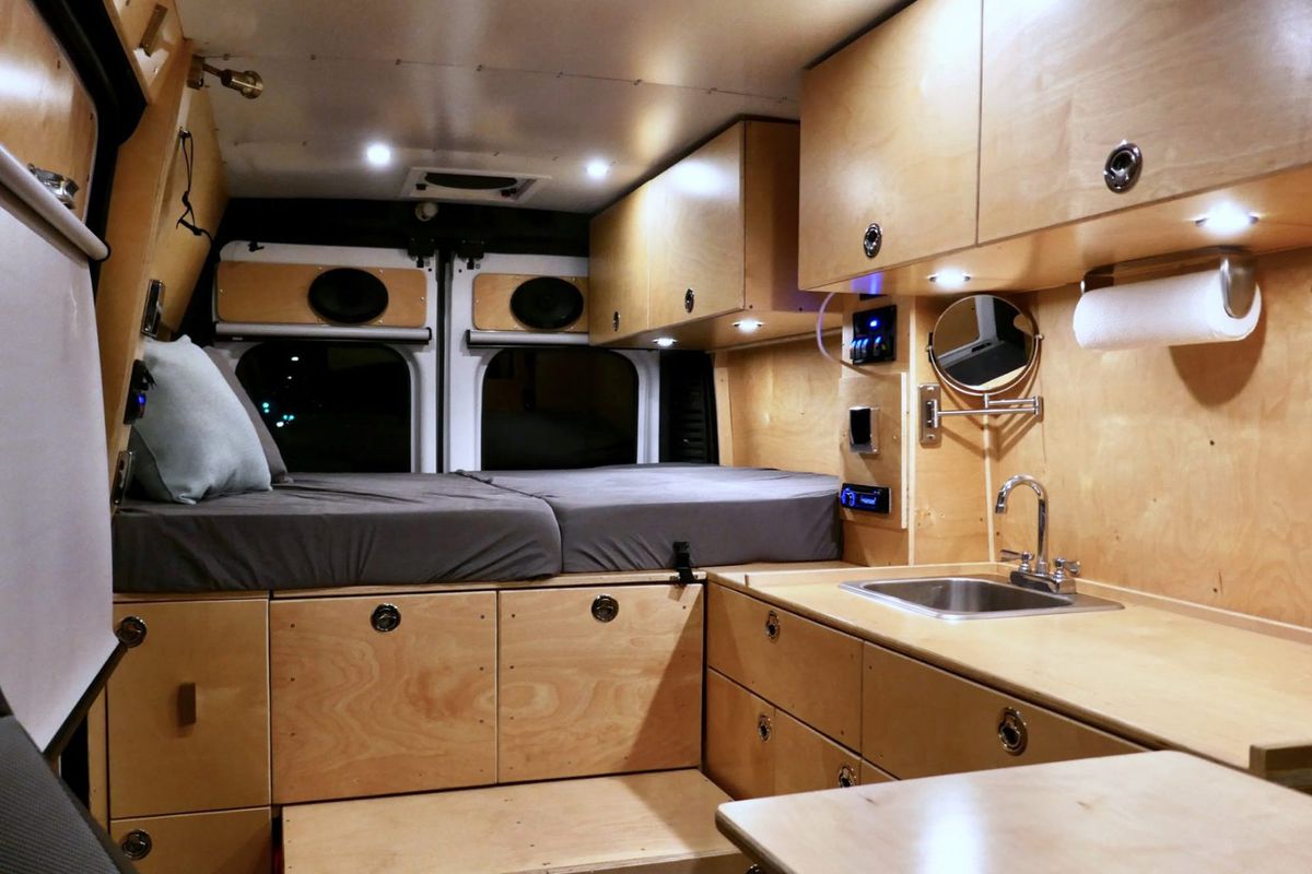 The interior of a camper van. The walls, roof, and cabinetry are wood. There is a bed with grey cushions and multiple pillows. There is a kitchenette with a sink.