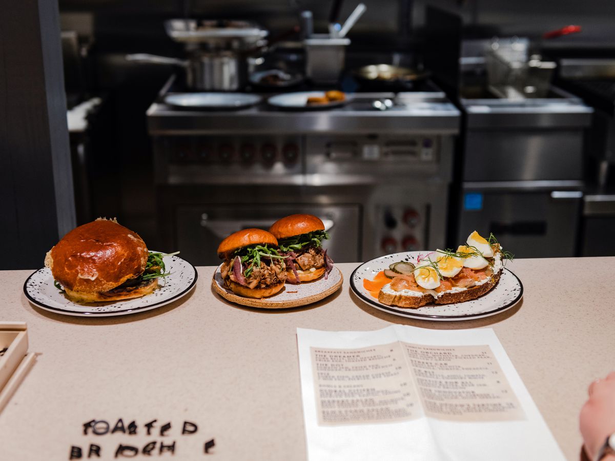 Three dishes lined up on a kitchen pass, ready to go out, with papers and cafe menu letters resting on the pass in the foreground.