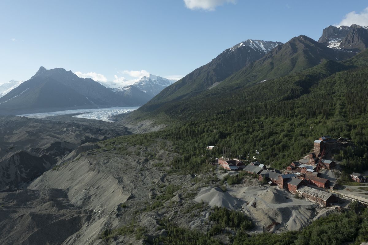 A sweeping image of a glacier and mountains, with crude rusty structures in the foregeground where a mine used to be. 