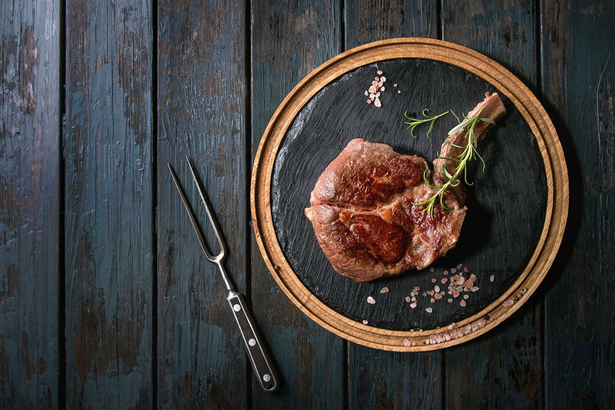 Grilled black angus beef tomahawk steak on bone served with salt, pepper and rosemary on round wooden slate cutting board with meat fork over dark wooden plank background Top view, copy space