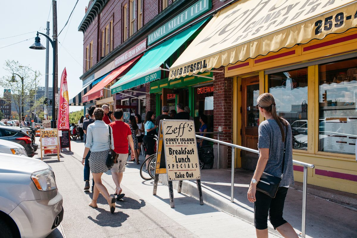 People walk past the yellow awning and sandwich board sign for Zeff’s on a sunny day in Eastern Market.