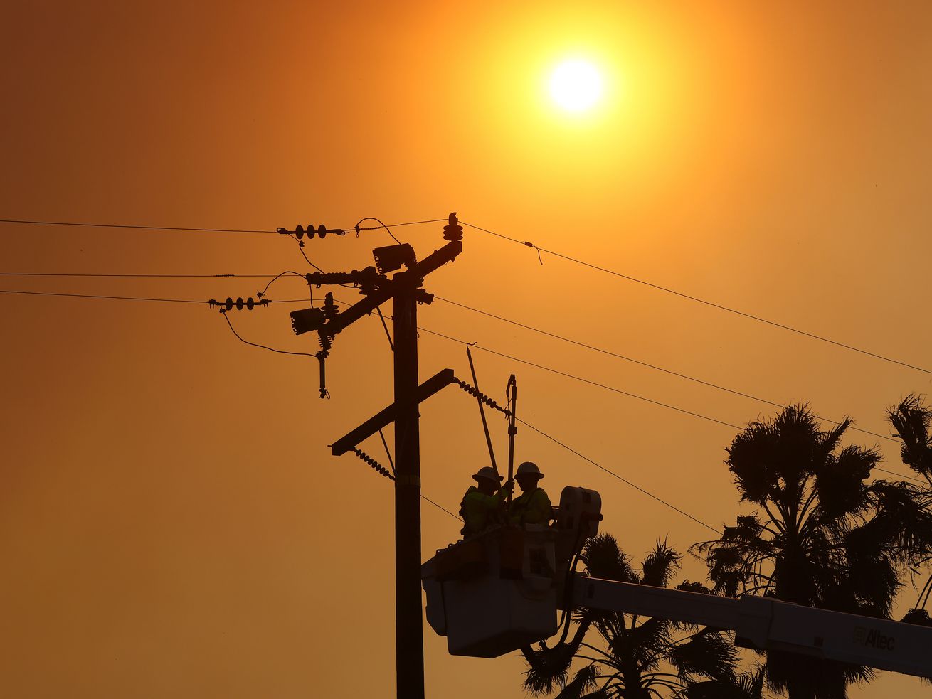 Workers in a cherry-picker basket approach a power pole while a bright sun glares overhead.