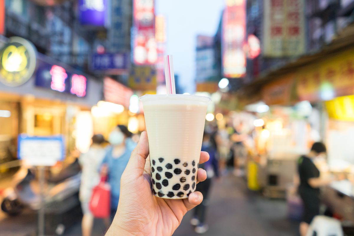 A hand holding up bubble tea; in the background, the lights of a Taiwanese night market.