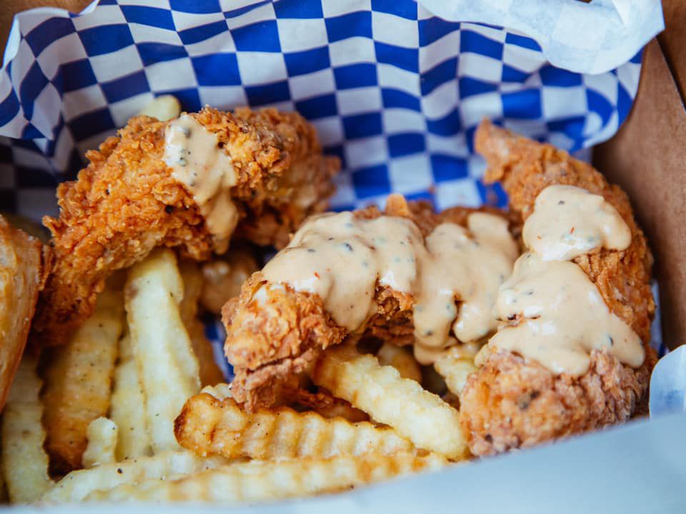 A box with white and blue checked paper filled with chicken fingers, covered in a creamy sauce, over crinkle cut fries