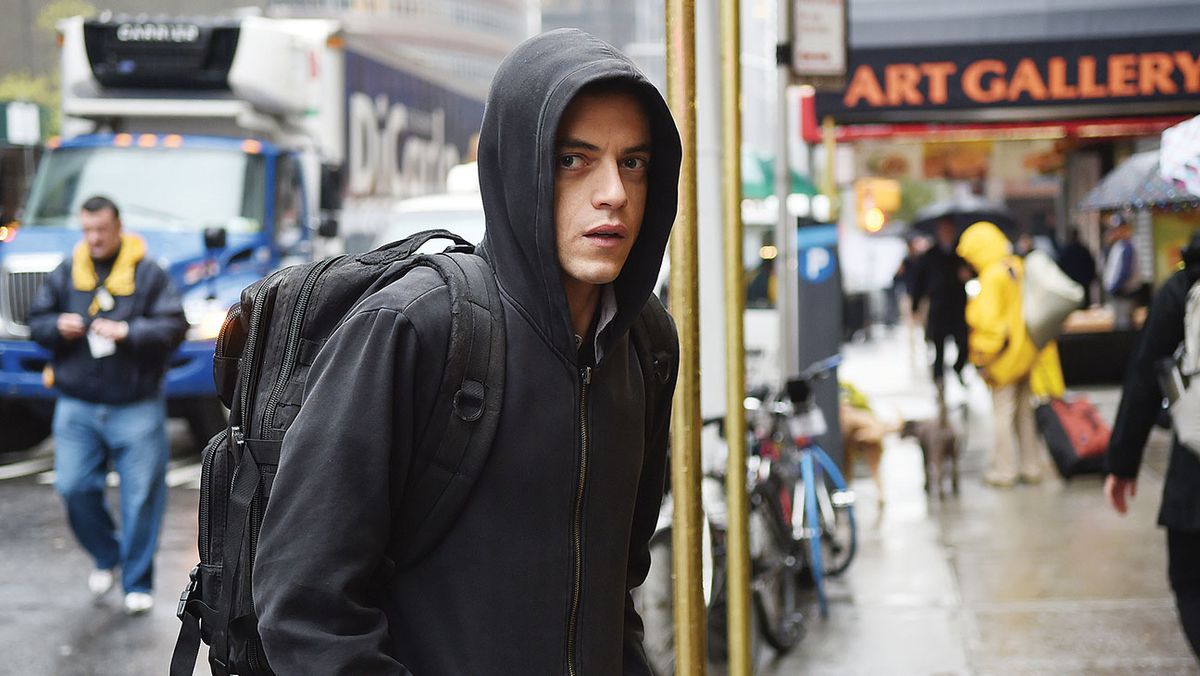 Rami Malek wears a black hoodie covering his head and a black backpack while walking on the street in Mr. Robot