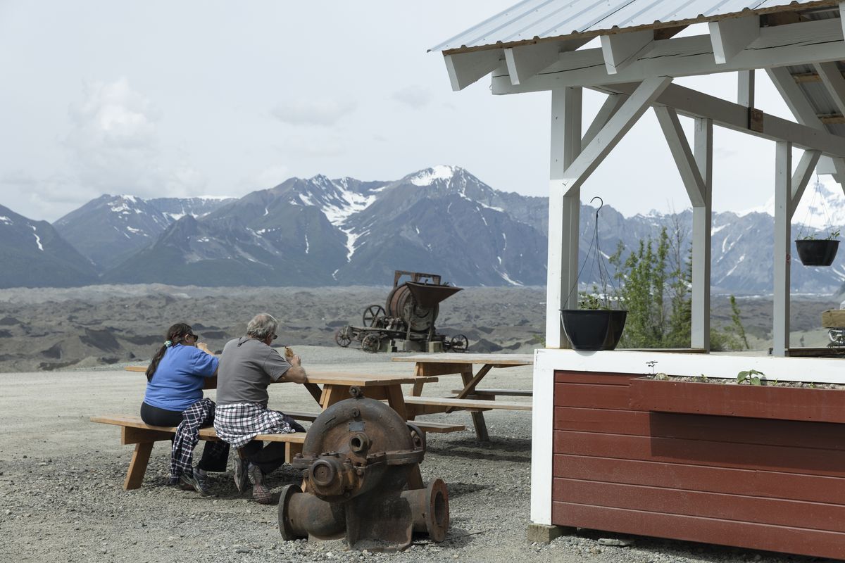 Two people sit on a picnic table in front of a red and white wooden structure, with mountains in the backround.