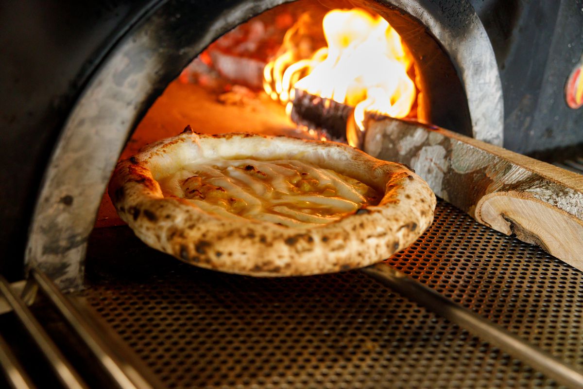 Pizza on a metal paddle going into an oven lit by large flames. 