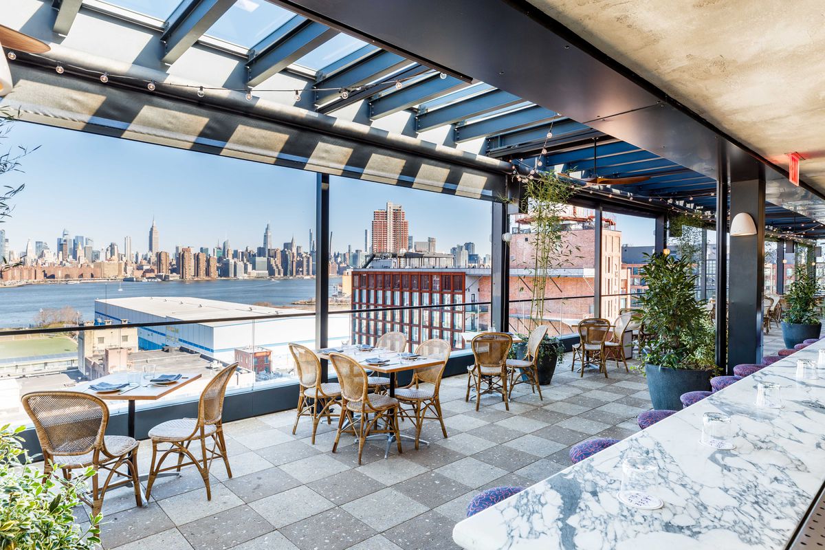 A rooftop, open-air dining room with light wooden tables and wicker chairs and a long bar, with a panoramic view of the Manhattan skyline